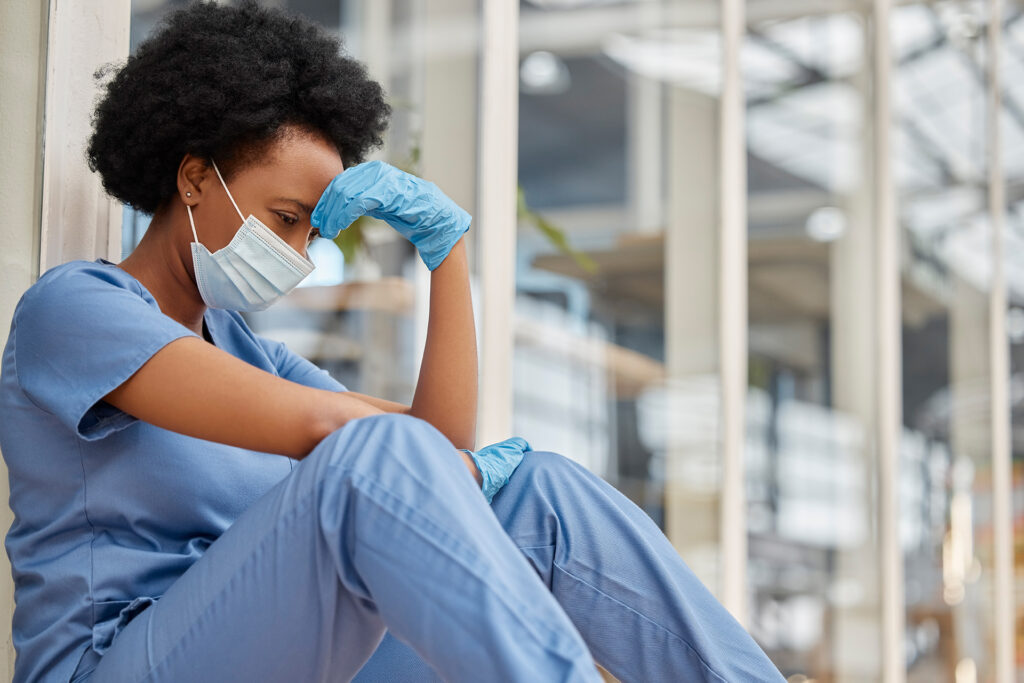 a nurse in scrubs and a facemask, sitting on the floor, her head on her hand, looking exhausted