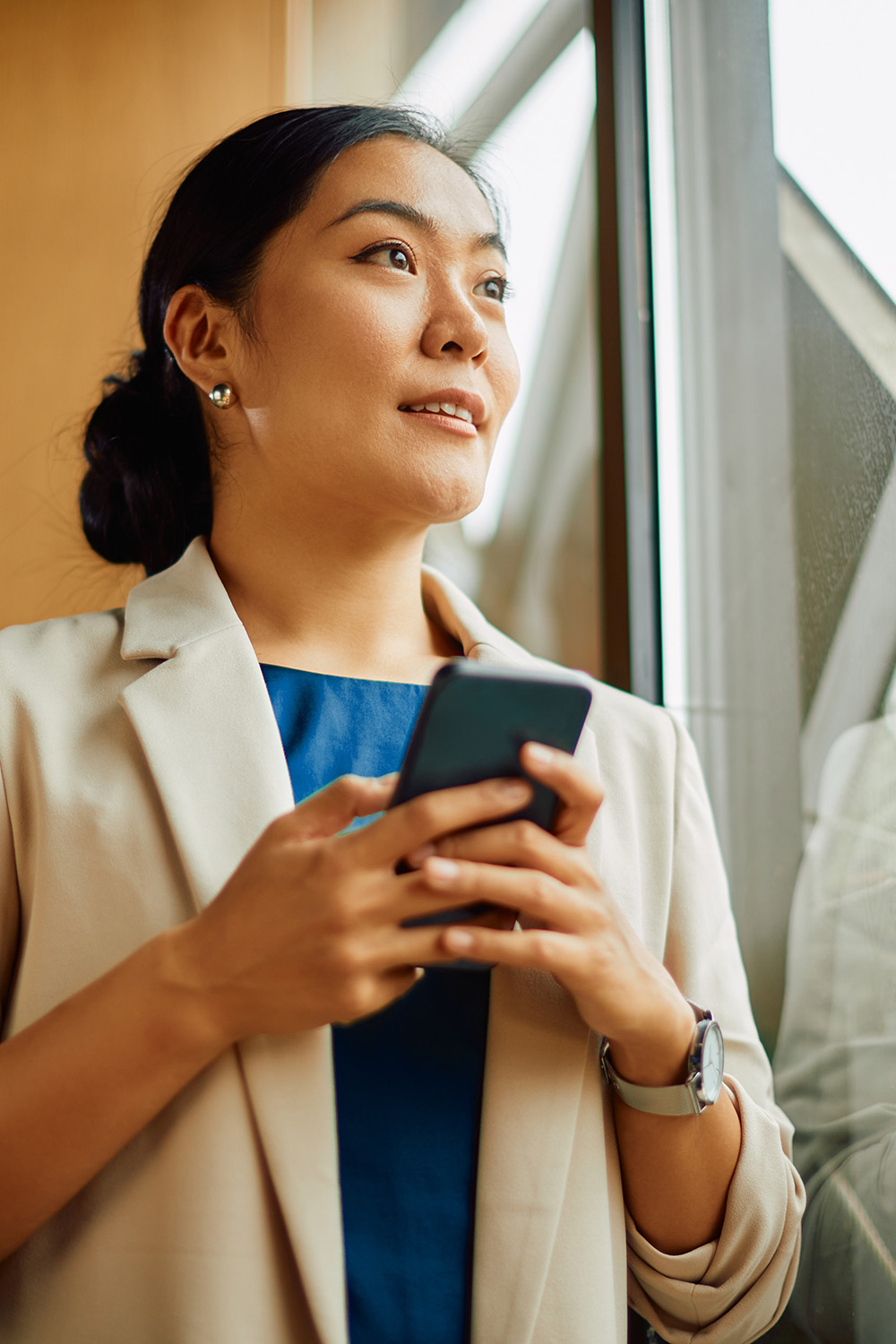 Young Asian businesswoman text messaging on mobile phone while standing by the window in the office and looking through it.