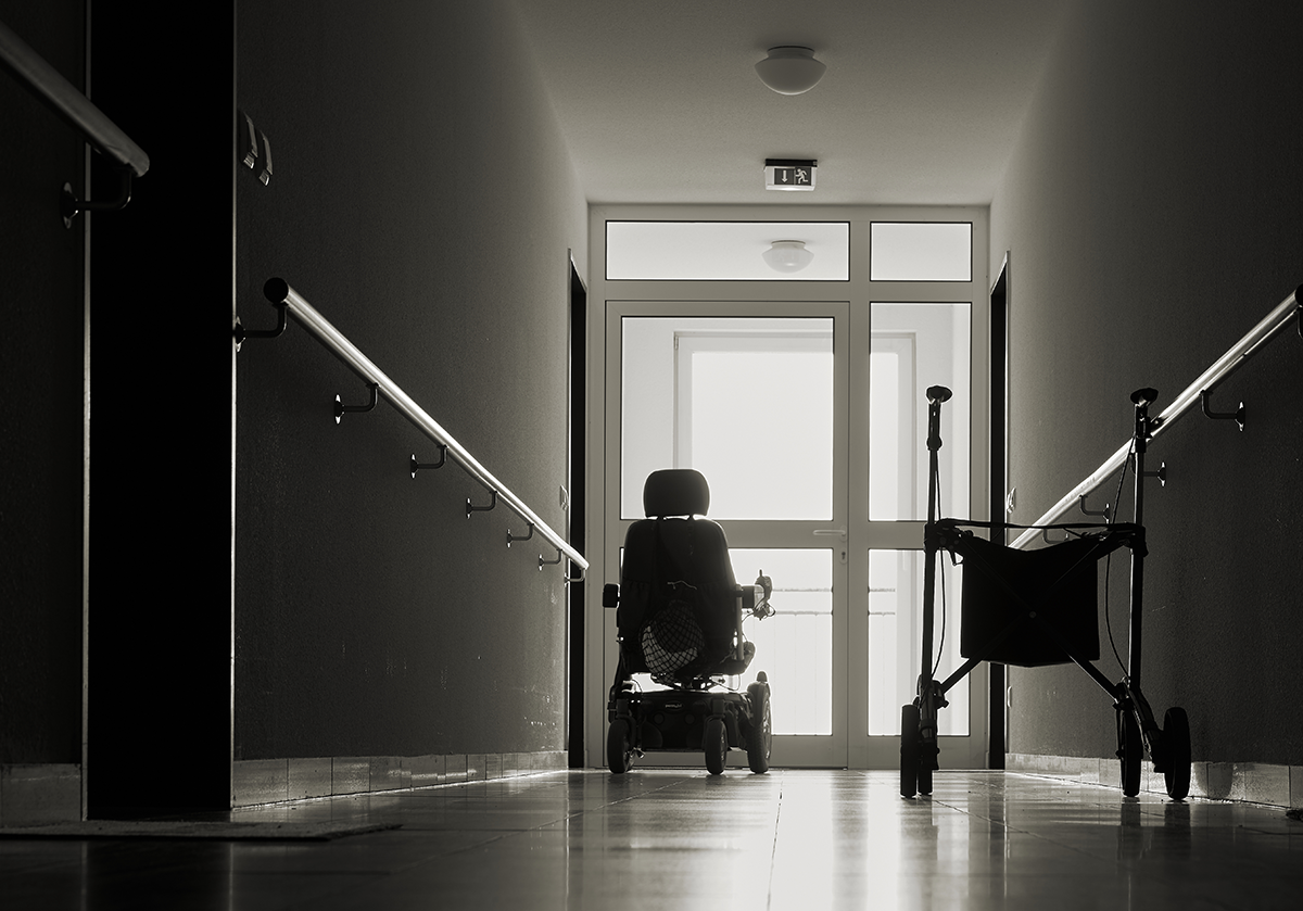 a black and white photo of a nursing home hallway showing an empty wheelchair and an exit sign